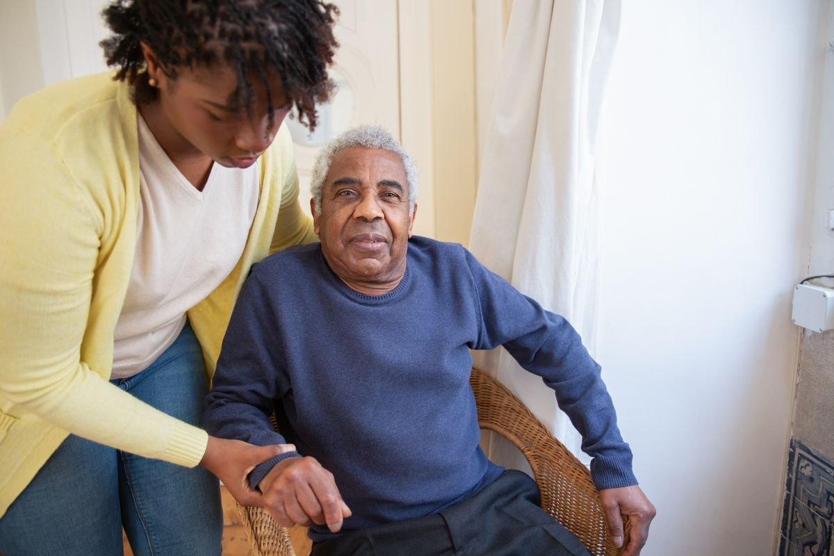 A caregiver sits beside an older man in his living room, resting a hand on his arm as they talk.