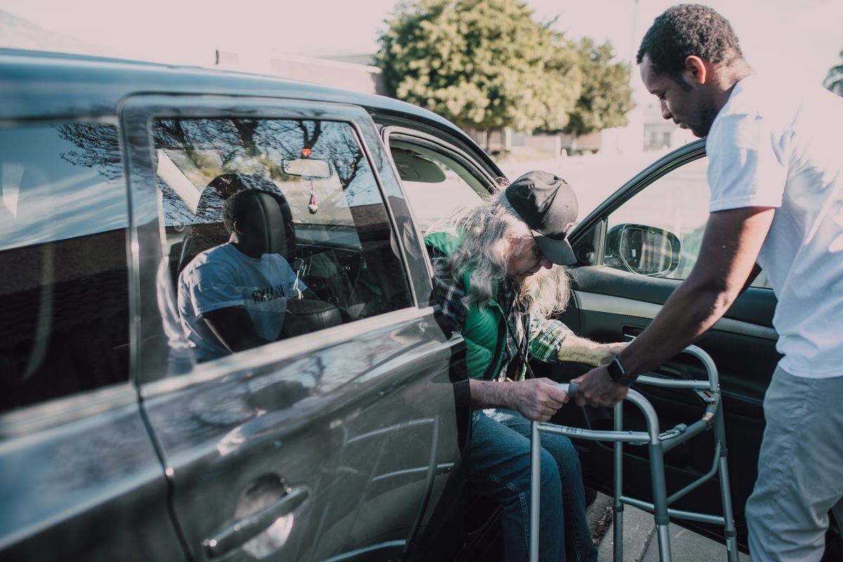 An attendant supports an older passenger with a walker as they step into a waiting vehicle.