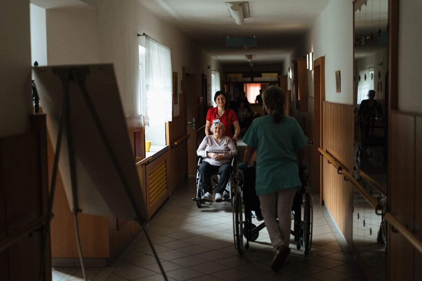 Two staff members push patients in wheelchairs along a bright hospital corridor.