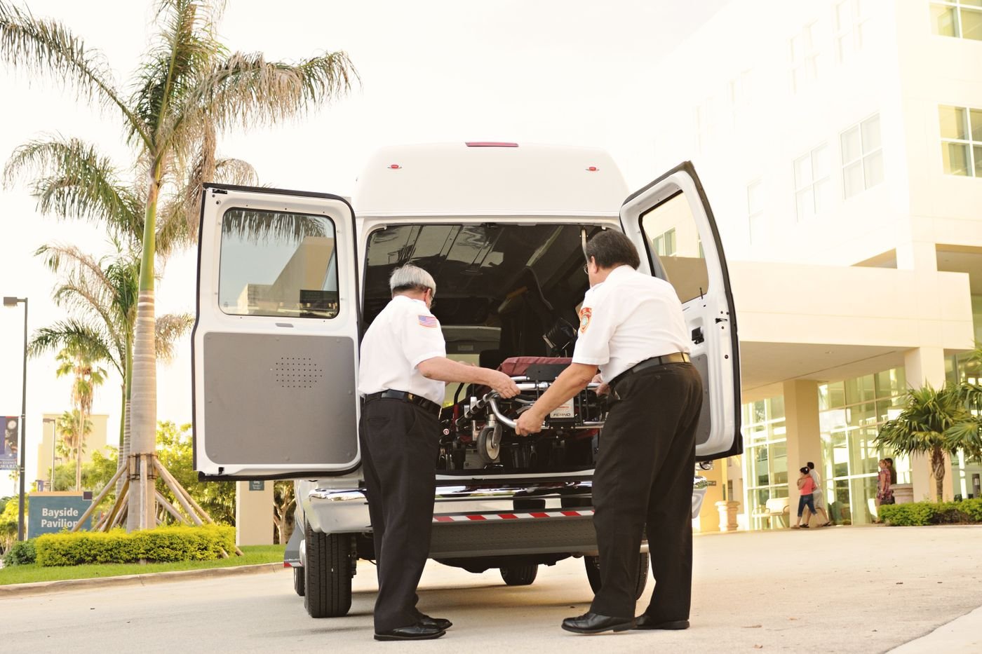 Two attendants secure a stretcher inside the rear of a patient transfer van parked at a hospital entrance.