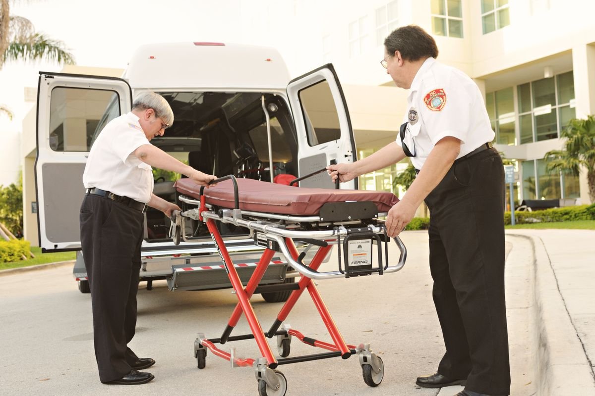 Two uniformed attendants guide a made-up stretcher toward the rear doors of a patient transfer van.