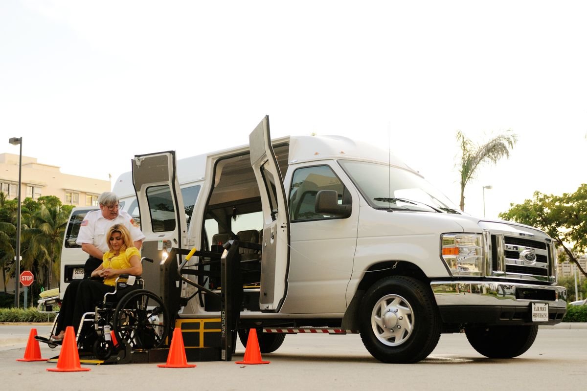 A passenger seated in a wheelchair is guided onto the ramp of an accessible transfer van by an attendant.