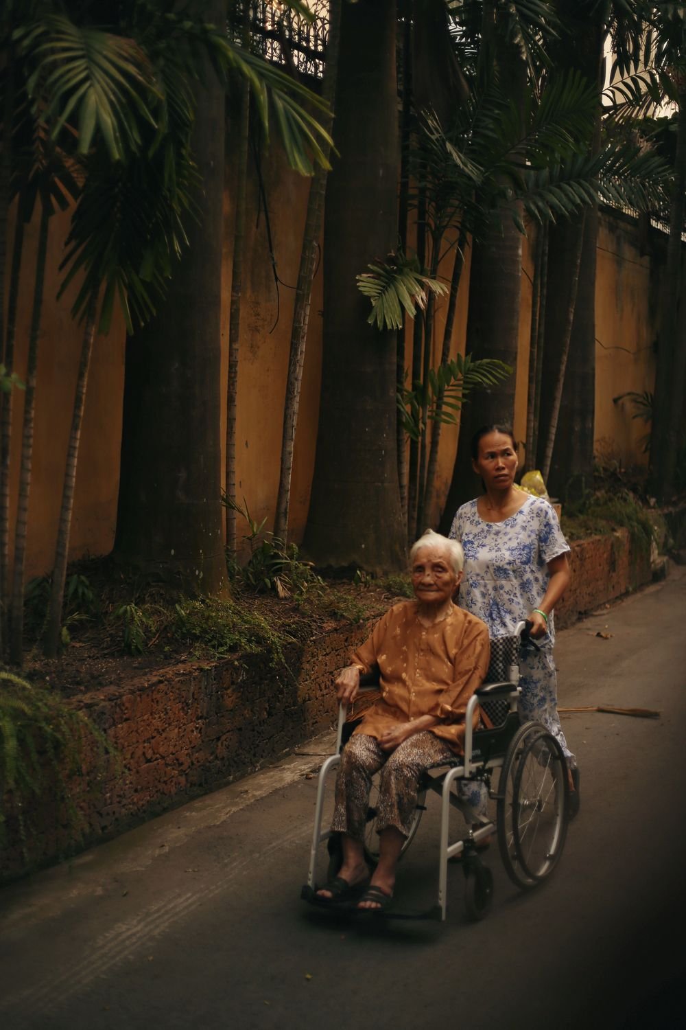 A care worker walks alongside an older woman she is pushing in a wheelchair on a quiet residential street.